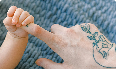 Baby hand wrapped around an adult’s tattooed finger against a blue‑gray knitted background.